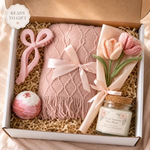 Könnte beinhalten: Eine Geschenkbox mit einer rosa Strickdecke mit Satinband, einer rosa Haarspange, einer runden rosa-weißen Badebombe, einem Strauß aus drei gehäkelten Tulpen in Rosa und Pfirsich sowie einer Kerze mit dem Etikett To an Amazing New Mom.
