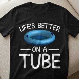 Könnte beinhalten: Dunkelgraues T-Shirt mit dem weißen Text "LIFE'S BETTER ON A TUBE". Eine blaue Schwimmreifen-Grafik ist über dem Text zentriert. Das Shirt besteht aus einem weichen Material.