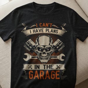 Könnte beinhalten: Dunkelgraues T-Shirt mit einem Grafikdesign mit einem Totenkopf, Kolben und Schraubenschlüsseln. Der Text lautet "I CAN'T I HAVE PLANS IN THE GARAGE" in Orange und Weiß. Das Shirt hat einen Rundhalsausschnitt und kurze Ärmel.