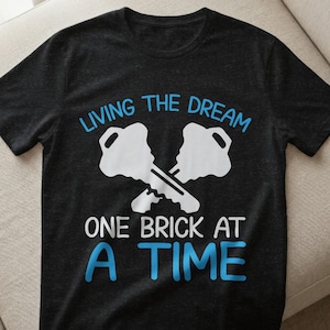 Könnte beinhalten: Dunkelgraues T-Shirt mit dem Text "LIVING THE DREAM ONE BRICK AT A TIME" in Blau und Weiß. Das Design beinhaltet zwei gekreuzte Schlüssel in Weiß.