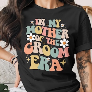 Könnte beinhalten: Schwarzes T-Shirt mit dem Text "IN MY MOTHER OF THE GROOM ERA" in einem Retro-Stil. Der Text ist in verschiedenen Farben wie Orange, Gelb, Blau und Rosa gehalten, mit Gänseblümchen-Akzenten. Ein festliches Design.