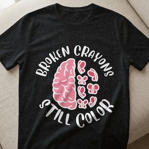 May include: Dark gray t-shirt with the text "BROKEN CRAYONS STILL COLOR" in white. The design features a pink brain and pink butterfly shapes. The shirt is made of a soft material.