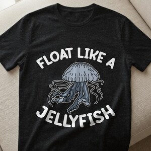 Könnte beinhalten: Dunkelgraues T-Shirt mit einer Illustration einer Qualle in Grau- und Blautönen. Der Text "FLOAT LIKE A JELLYFISH" ist in Weiß über und unter dem Quallen-Design gedruckt, was ein maritimes Thema erzeugt.