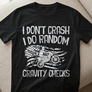 以下が含まれることがあります： 白のテキストとグラフィックが特徴の黒いTシャツ。テキストは「I DON'T CRASH I DO RANDOM GRAVITY CHECKS」と書かれています。グラフィックは、アメリカ国旗のデザインのオートバイに乗ったモトクロスライダーを描いています。