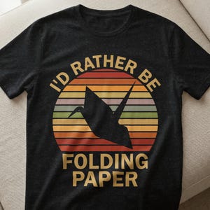Könnte beinhalten: Dunkelgraues T-Shirt mit einem kreisförmigen Design, das einen schwarzen Origami-Kranich vor einem Hintergrund in Sonnenuntergangsfarben zeigt. Der Text lautet "I'D RATHER BE FOLDING PAPER" in Gold.