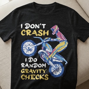 Könnte beinhalten: Schwarzes T-Shirt mit einem Grafikdesign eines Motorradfahrers, der einen Stunt ausführt. Der Text "I DON'T CRASH I DO RANDOM GRAVITY CHECKS" ist in Weiß und Gelb gehalten. Das Motorrad und der Fahrer sind blau, gelb und rot.