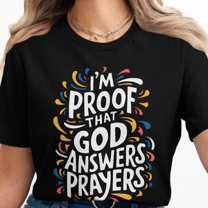 Könnte beinhalten: Schwarzes T-Shirt mit dem weißen Text "I'M PROOF THAT GOD ANSWERS PRAYERS", umgeben von bunten Wirbeln in Blau, Gelb und Rot. Das T-Shirt hat einen Rundhalsausschnitt.