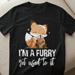 May include: Dark gray t-shirt featuring a cartoon fox with brown and white fur, holding a bubble tea. The text "I'M A FURRY get used to it" is printed in white. The fox is smiling, and there are two small pink hearts.
