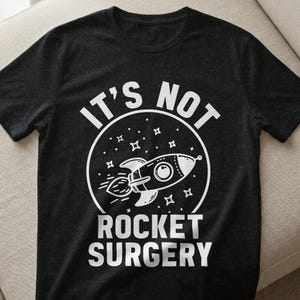Könnte beinhalten: Dunkelgraues T-Shirt mit einem weißen Grafikdesign einer Rakete in einem Kreis mit Sternen. Der Text "IT'S NOT ROCKET SURGERY" ist über und unter dem Raketendesign gedruckt.