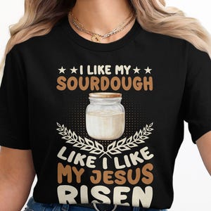 Könnte beinhalten: Schwarzes T-Shirt mit dem Text "I LIKE MY SOURDOUGH LIKE I LIKE MY JESUS RISEN." Das Design umfasst ein Glas Sauerteig und Weizenähren. Der Text ist braun und weiß.