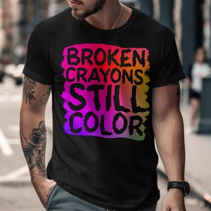 Könnte beinhalten: Schwarzes T-Shirt mit dem Text "BROKEN CRAYONS STILL COLOR" in einem farbenfrohen, mehrfarbigen Design. Der Text ist in einem rechteckigen Rahmen platziert. Das T-Shirt hat kurze Ärmel und ist aus weichem Stoff.