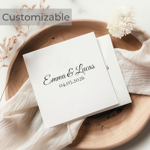 Könnte beinhalten: Zwei weiße quadratische Karten mit den Namen "Emma & Lucas" und dem Datum "04.05.2026" in schwarzer Schrift gedruckt. Die Karten liegen auf einer Holzschale mit weißem Stoff und Trockenblumen.