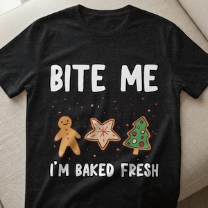 Könnte beinhalten: Schwarzes T-Shirt mit dem Text "BITE ME" über Lebkuchenmännchen, Stern- und Weihnachtsbaum-Keksen. Darunter steht "I'M BAKED FRESH". Die Kekse sind braun, rot und grün, passend zur Weihnachtszeit.