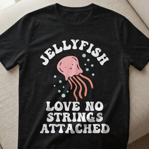 Könnte beinhalten: Schwarzes T-Shirt mit einer rosa Quallen-Illustration und dem Text "JELLYFISH" darüber und "LOVE NO STRINGS ATTACHED" darunter. Die Qualle hat ein lächelndes Gesicht und orangefarbene Tentakel.