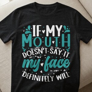 Può includere: T-shirt nero con la scritta "IF MY MOUTH DOESN'T SAY IT my face DEFINITELY WILL" in bianco e verde acqua. Il design include cuori ed elementi decorativi.
