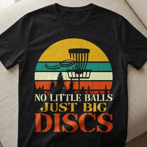 May include: Dark grey t-shirt featuring a disc golf design with a basket, discs, and trees against a sunset backdrop. The text reads "NO LITTLE BALLS JUST BIG DISCS" in white and orange.