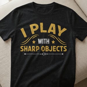 Könnte beinhalten: Schwarzes T-Shirt mit dem Text "I PLAY WITH SHARP OBJECTS" in verblichener goldener Schrift. Der Text ist in zwei Zeilen mit dekorativen Elementen angeordnet. Ein lässiges, lustiges T-Shirt.