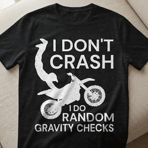 以下が含まれることがあります： 黒いTシャツに、モトクロスバイクの白いグラフィックと「I DON'T CRASH I DO RANDOM GRAVITY CHECKS」の文字。デザインは空中のライダーを特徴とし、アクション満載のテーマに貢献しています。