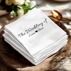 Könnte beinhalten: Ein Stapel weißer Servietten mit dem schwarzen Text "The Wedding of Emma ❤ Lucas". Die Servietten liegen auf einer Holzschale, mit einem verschwommenen Hintergrund aus Blumen und einem Löffel.