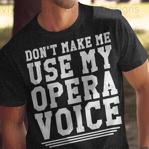 Könnte beinhalten: Dunkelgraues T-Shirt mit dem Text "DON'T MAKE ME USE MY OPERA VOICE" in fetten weißen Buchstaben. Das Design beinhaltet drei horizontale Linien am unteren Rand. Das Shirt hat einen Rundhalsausschnitt und scheint aus weichem Material zu sein.