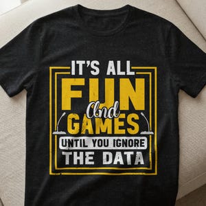 Könnte beinhalten: Schwarzes T-Shirt mit gelb-weißem Aufdruck. Der Aufdruck lautet: "IT'S ALL FUN and GAMES UNTIL YOU IGNORE THE DATA". Der Text ist in einem gelben Rechteck eingerahmt. Das T-Shirt ist aus Baumwolle.