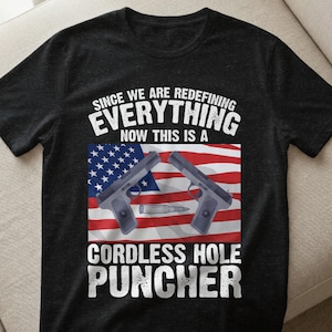 Könnte beinhalten: Dunkelgraues T-Shirt mit einem grafischen Design. Das Design zeigt zwei graue Pistolen, eine Kugel und eine amerikanische Flagge. Der Text lautet: "SINCE WE ARE REDEFINING EVERYTHING NOW THIS IS A CORDLESS HOLE PUNCHER."