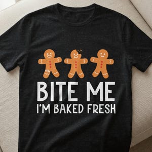 Könnte beinhalten: Dunkelgraues T-Shirt mit drei Lebkuchenmännern mit orangefarbener Glasur und weißen Zuckergussdetails. Der Text "BITE ME" und "I'M BAKED FRESH" ist in Weiß gedruckt.