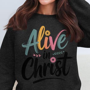 Könnte beinhalten: Dunkelgraues Sweatshirt mit dem Schriftzug "Alive in Christ" in farbenfroher Schrift. Das Wort "Alive" ist in Türkis, Gelb und Rosa gehalten, mit floralen Akzenten. Das Wort "Christ" ist in einer kräftigen schwarzen Schrift geschrieben.