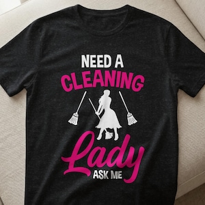 Könnte beinhalten: Schwarzes T-Shirt mit dem Text "NEED A CLEANING Lady ASK ME" in Weiß und Pink. Das Design zeigt eine weiße Silhouette einer Person, die einen Besen hält, mit zwei Besen auf beiden Seiten.