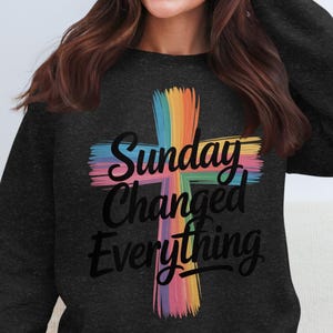 Könnte beinhalten: Dunkelgraues Sweatshirt mit einem farbenfrohen Kreuzdesign und dem Schriftzug "Sunday Changed Everything" in schwarzer Schrift. Das Kreuz ist in Regenbogenfarben gemalt und verleiht der Kleidung eine lebendige Note.