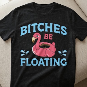 Könnte beinhalten: Schwarzes T-Shirt mit dem Schriftzug "BITCHES BE FLOATING" in Hellblau, mit einem pinkfarbenen Flamingo-Schwimmring. Der Flamingo hat einen braunen Schnabel und ist von blauen Wasserspritzern umgeben. Das Shirt ist aus weichem Material.
