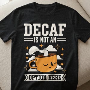 Könnte beinhalten: Dunkelgraues T-Shirt mit dem weißen Text "DECAF IS NOT AN OPTION HERE". Darunter befindet sich eine Cartoon-Kaffeetasse mit Dampf und Sternen.
