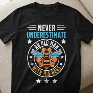 Könnte beinhalten: Schwarzes T-Shirt mit dem Text "NEVER UNDERESTIMATE AN OLD MAN WITH HIS BEES". Das Design zeigt eine Biene mit blauen Flügeln und einem gestreiften Körper, umgeben von Sternen. Ein lässiges Kleidungsstück.