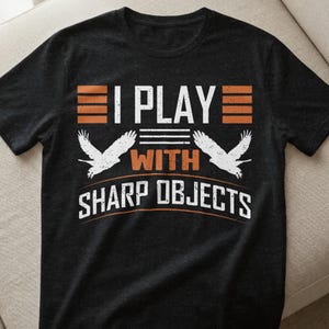 Könnte beinhalten: Schwarzes T-Shirt mit dem weißen Text "I PLAY WITH SHARP OBJECTS" und orangefarbenen Akzenten. Das Design umfasst zwei weiße Adlersilhouetten. Ein lässiges Grafik-T-Shirt.