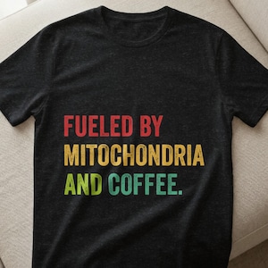 Könnte beinhalten: Schwarzes T-Shirt mit dem Text "FUELED BY MITOCHONDRIA AND COFFEE" in roten, gelben und grünen Blockbuchstaben. Das T-Shirt hat einen Rundhalsausschnitt und kurze Ärmel. Das T-Shirt ist aus weichem Material.