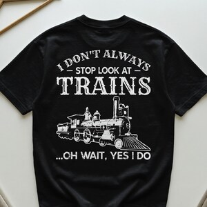 Könnte beinhalten: Schwarzes T-Shirt mit weißer Grafik einer Dampflokomotive und dem Text "I DON'T ALWAYS - STOP LOOK AT - TRAINS ...OH WAIT, YES I DO". Das Design zeigt eine detaillierte Illustration einer klassischen Lokomotive.