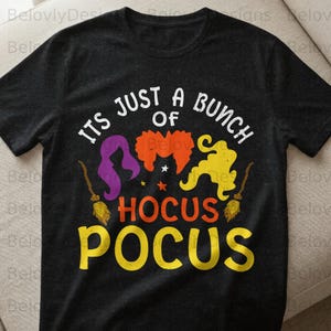 Könnte beinhalten: Dunkelgraues T-Shirt mit dem Text "ITS JUST A BUNCH OF HOCUS POCUS" in Weiß, Orange und Gelb. Das Design umfasst drei Hexensilhouetten in Lila, Orange und Gelb mit zwei Besen.