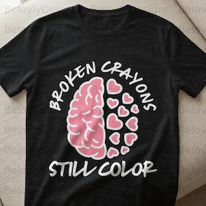 May include: Dark gray t-shirt with the text "BROKEN CRAYONS STILL COLOR" in white. The design features a pink brain and a collection of pink hearts. The shirt is a crew neck.