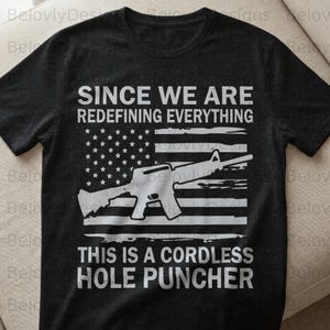 Könnte beinhalten: Dunkelgraues T-Shirt mit weißer Grafik einer amerikanischen Flagge mit Gewehr. Der Text lautet "SINCE WE ARE REDEFINING EVERYTHING" und "THIS IS A CORDLESS HOLE PUNCHER."