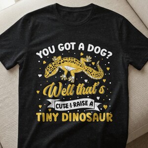 Könnte beinhalten: Schwarzes T-Shirt mit einer gelb-schwarzen Gecko-Illustration und dem Text "YOU GOT A DOG? Well that's CUTE I RAISE A TINY DINOSAUR". Das Design enthält Herzen und dekorative Elemente.