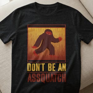 Könnte beinhalten: Dunkelgraues T-Shirt mit einem Bigfoot-Motiv in einem rechteckigen Rahmen. Die Illustration zeigt einen braunen Bigfoot, der mit roten Händen und Füßen vor einem gelb-orangen Hintergrund geht. Der Text "DON'T BE AN ASSQUATCH" steht darunter.