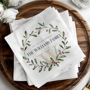 Könnte beinhalten: Ein Stapel weißer Papierservietten mit einem aufgedruckten Kranz aus grüner Mistel und roten Beeren. Der Text lautet JOY TO THE WORLD, THE WILLIAMS FAMILY und 2026, zentriert um ein großes goldenes W mit feinem Streifenmuster.