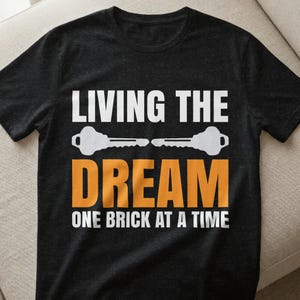 Könnte beinhalten: Schwarzes T-Shirt mit dem Text "LIVING THE DREAM" in Weiß, mit Schlüsselillustrationen. Das Wort "DREAM" ist orange, und der Satz "ONE BRICK AT A TIME" ist weiß. Das Shirt besteht aus einem weichen Material.