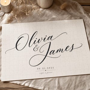 Könnte beinhalten: Ein rechteckiges Puzzle mit cremefarbener Oberfläche, das den schwarzen Schriftzug Olivia & James, das Datum 21.06.2025, ein kleines Herzsymbol und den Text sign a piece in der Mitte der ineinandergreifenden Puzzleteile zeigt.