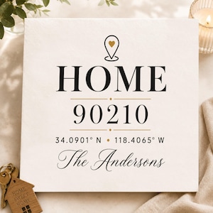 May include: A square white sign featuring the word HOME in black capital letters with a location pin icon containing a small gold heart above it. Below are the numbers 90210, geographic coordinates, and the text The Andersons in black script.