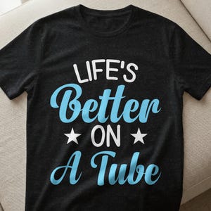 Könnte beinhalten: Schwarzes T-Shirt mit dem Schriftzug "LIFE'S Better ON A Tube" in Weiß und Hellblau. Zwei weiße Sterne flankieren das Wort "ON". Das Shirt besteht aus einem weichen Material.