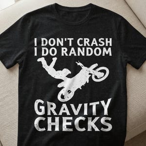Könnte beinhalten: Dunkelgraues T-Shirt mit weißem Text und Grafik. Der Text lautet „I DON'T CRASH I DO RANDOM GRAVITY CHECKS“. Die Grafik ist eine Silhouette einer Person und eines Motorrads in der Luft.