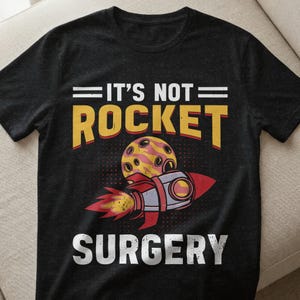 Könnte beinhalten: Schwarzes T-Shirt mit dem Text "IT'S NOT ROCKET SURGERY" in Weiß, Gelb und Rot. Das Design zeigt eine Rakete und einen planetenähnlichen Gegenstand, was eine humorvolle Grafik ergibt.