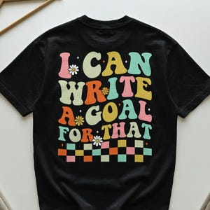 Könnte beinhalten: Schwarzes T-Shirt mit dem farbenfrohen Retro-Schriftzug "I CAN WRITE A GOAL FOR THAT". Das Design beinhaltet Gänseblümchen und ein Schachbrettmuster. Ein lässiges und motivierendes Grafik-T-Shirt.