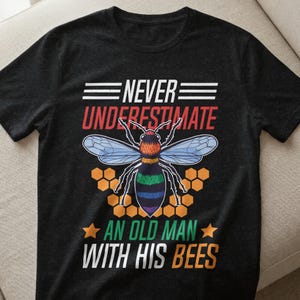 Könnte beinhalten: Schwarzes T-Shirt mit einem farbenfrohen Bienen-Design und dem Text "NEVER UNDERESTIMATE AN OLD MAN WITH HIS BEES". Die Biene hat blaue Flügel und einen gestreiften Bauch, umgeben von Waben.
